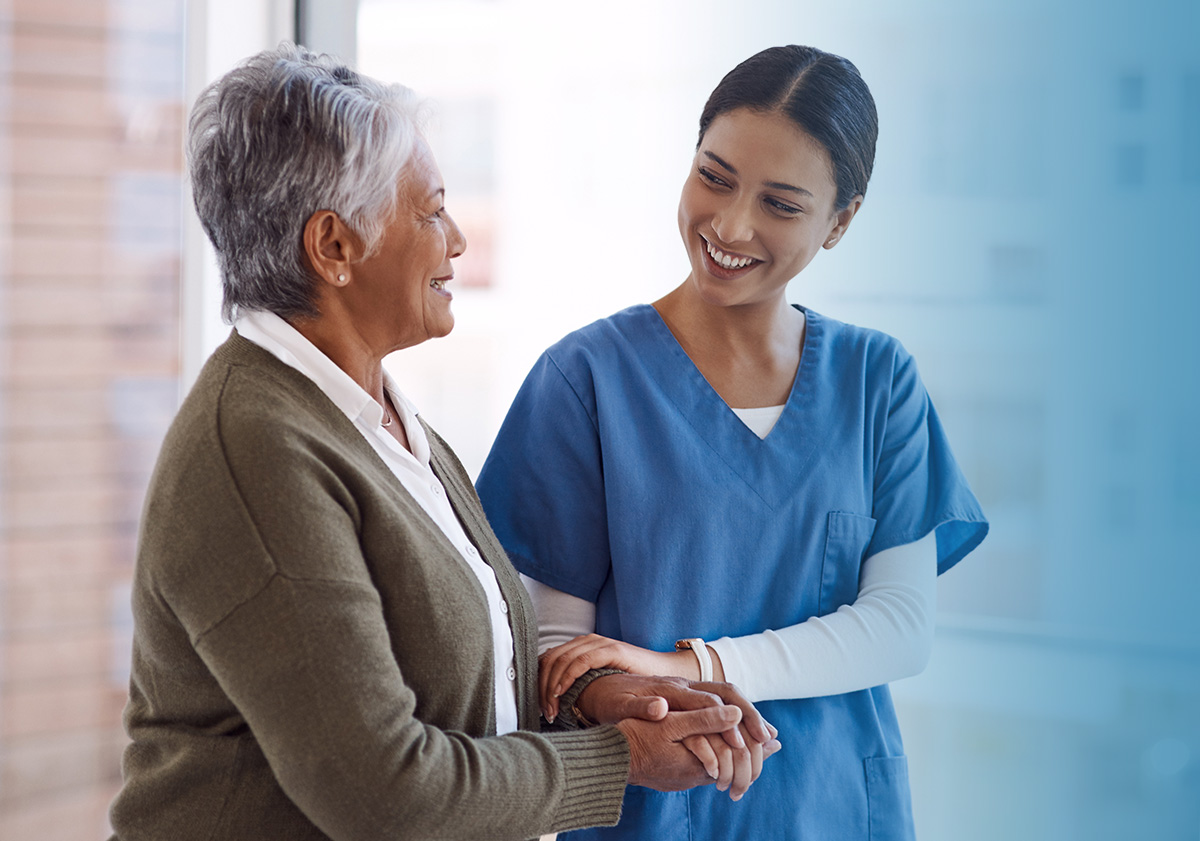 A nurse in blue scrubs holding the hands of an older woman patient, both smiling